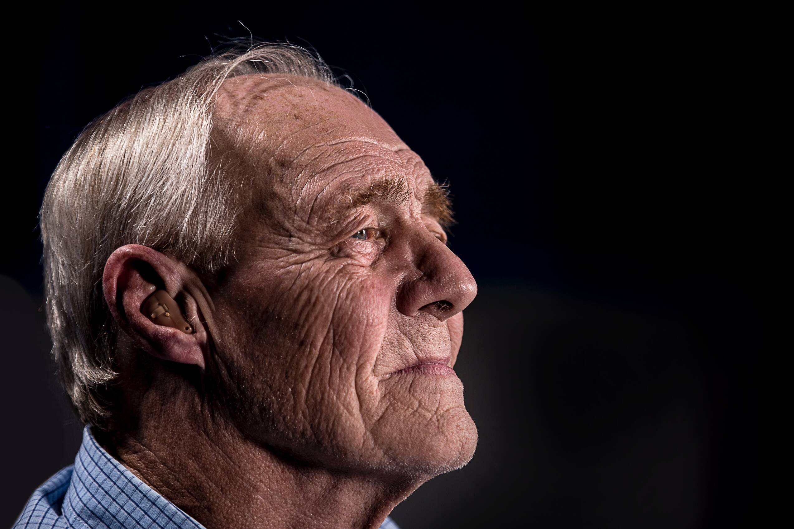Old man with wrinkle face and gray hair looking out and using a hearing aid.